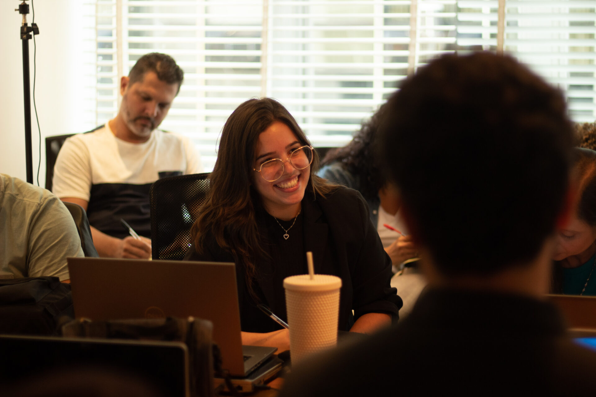 Uma mulher de pele clara com cabelos longos de cor castanho escuro utilizando roupas pretas e um óculos com hastes douradas, em frente à uma mesa cheia de laptops sorrindo e praticando networking em uma reunião empresarial com algumas pessoas em volta.